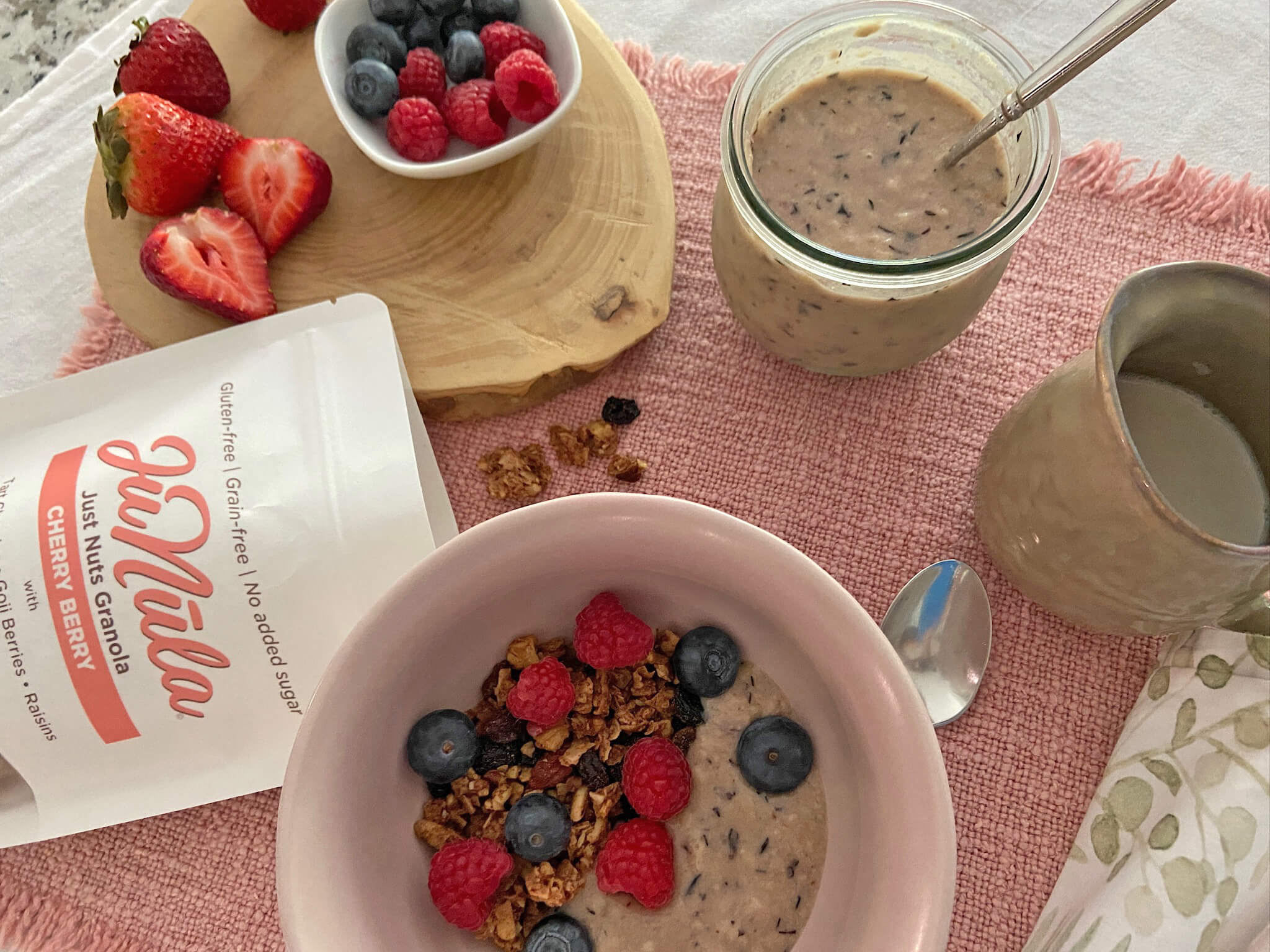 Breakfast scene with a bowl of Cherry Berry granola, berries, and a jar of yogurt on a pink tablecloth.
