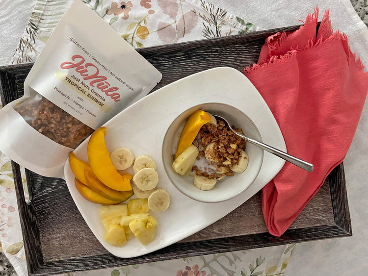 Bowl of Tropical Sunrise granola with bananas, mangoes, and pineapple on a tray next to a package of JuNūla granola.