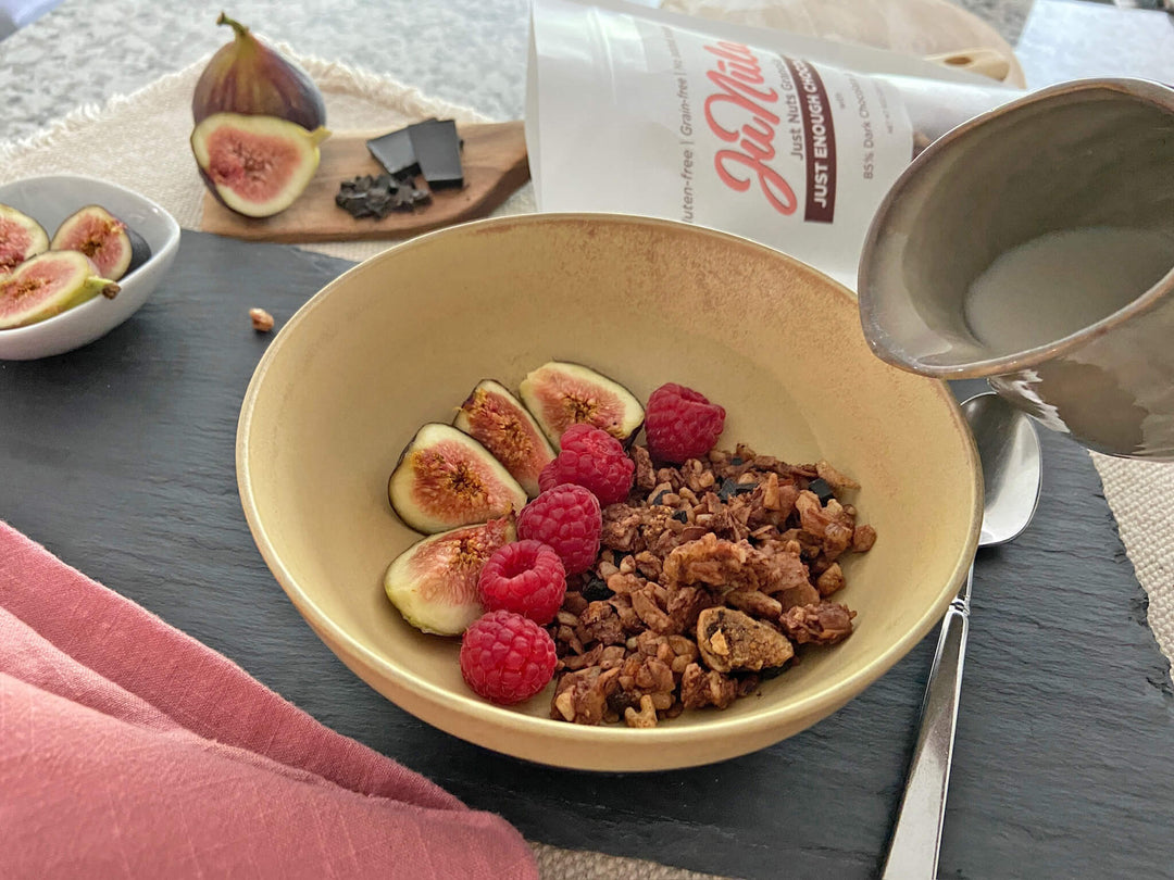 Bowl of Just Enough Chocolate granola with figs and raspberries on a slate surface, with a spoon and a container of yogurt in the background.