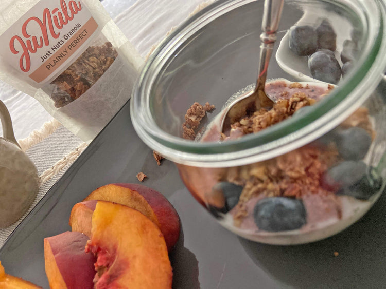 Glass jar with Plainly Perfect granola, blueberries, and a spoon on a table with a container of granola in the background.