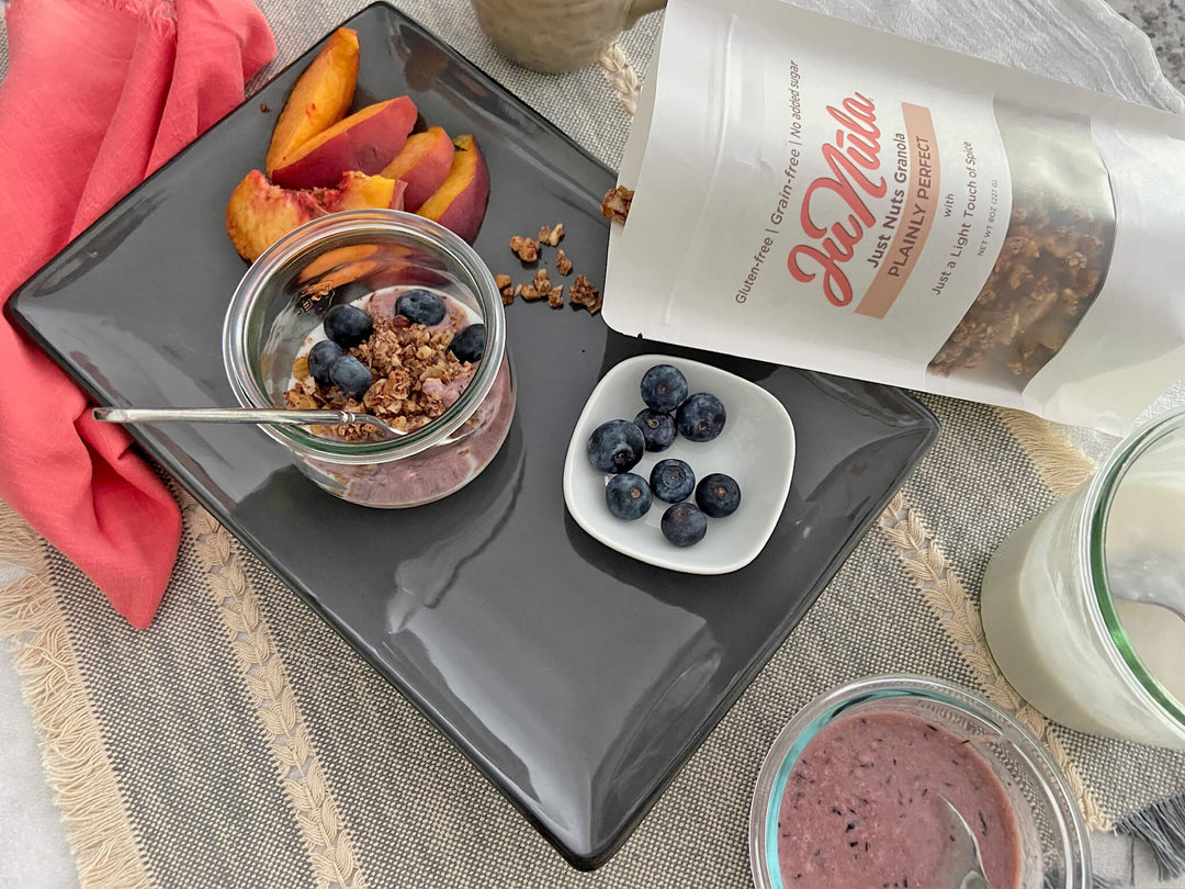 Yogurt parfait with Plainly Perfect granola, peaches, and blueberries on a dark plate with a container of granola.
