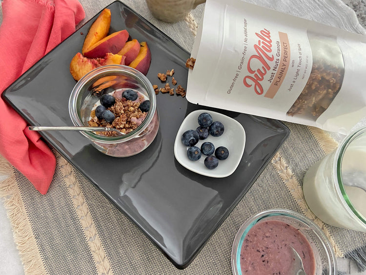 Yogurt parfait with Plainly Perfect granola, peaches, and blueberries on a dark plate with a container of granola.